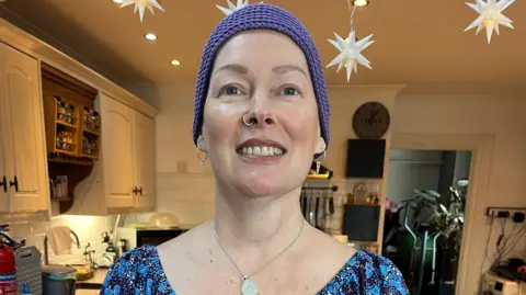 BBC A woman in a flowery blue dress, with a blue headscarf is standing in front of the camera holding her hands in front of her. She has a nose ring, and is wearing bangles and a watch on her wrists. She is smiling.