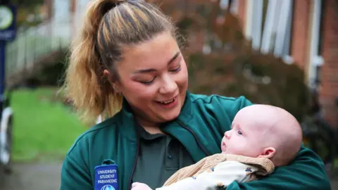 University of Worcester A student paramedic wit light brown hair tied in a ponytail. She is smiling down at a baby in her arms. She is wearing a teal t-shirt and jacket. On the jacket it reads 