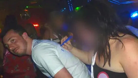 A photograph of Ian in a nightclub. A girl, with her face blurred to obscure her identity, writes on his white t-shirt with a blue marker pen.
