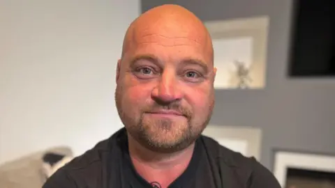 Offshore worker Phil Perry, who has a bald head and is wearing a black T-shirt, is smiling at the camera. He has tattoos on arms.