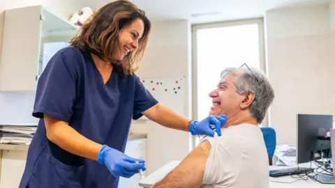 Getty Images Two people in a bland, white clinic room. One is an older man with grey hair, an incredibly intense grin on his face and his left arm is rolled up. A young woman with shoulder length brown hair, wearing a rich blue set of scrubs is standing over him. She holds a shot of flu vaccine in her medical gloved hands. 