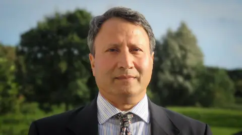 Rakesh Rakesh standing outside, trees in the background, the sun is shining. He is wearing a dark grey suit, yellow and blue striped shirt and a tie with elephant pattern