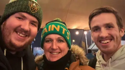 Family handout Three people - a smiling young man in green woollen hat on the left; a middle-aged woman in the centre in a green and yellow hat that says 