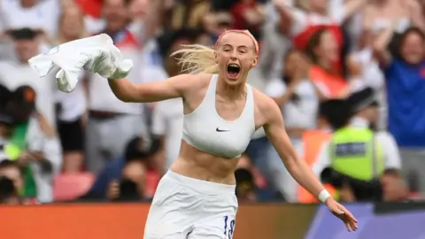 FRANCK FIFE/AFP via Getty Images A female footballer celebrates after scoring a goal. The player, who has blonde hair, is holding her white shirt above her head. She is wearing a white sports bra and white shorts. A crowd wearing red, white and blue clothing are visible in the background.