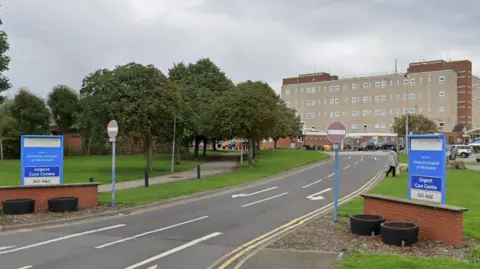 Google University Hospital of Hartlepool entrance with a road leading up to a large grey building. There are two blue signs on both sides of the road reading 'Urgent Care Centre' and 'No A&E'.