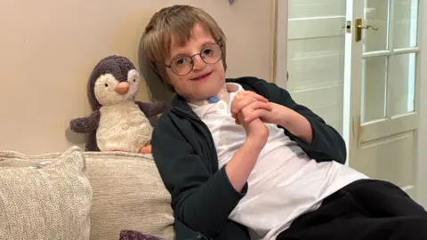 A boy with light-brown hair and black-rimmed glasses is leaning back on his beige sofa. He has a genetic condition and has a white plaster on his neck. He is wearing a white polo top and black fleece. He is clasping his hands together and smiling at the camera. Behind him is a grey and white toy penguin teddy. 