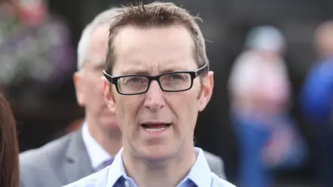 Pacemaker Philip McGuigan has short light brown hair and black-framed glasses and is speaking at an outdoor event