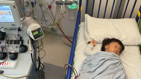 Oliver Chu lies in a hospital cot. He has his eyes closed and is under a pale blue blanket. Tubes are hooked up from a machine next to the cot.