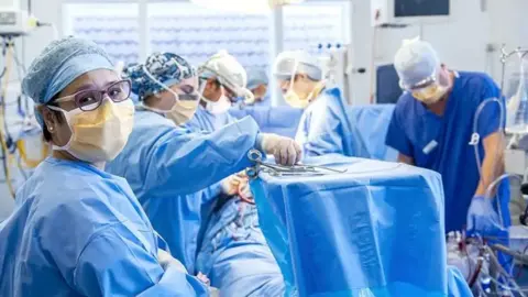 Royal Papworth A hospital theatre where surgery is being carried out. Five surgeons gather around an unseen patient, who is covered with blue paper cloths. The surgeons all wear matching blue gowns and wear masks and caps. On a high tray are surgical instruments, with one surgeon reaching for a scalpel.