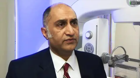 Amir Bhatti wearing a dark suit jacket, white shirt, and red striped tie standing indoors next to a large medical imaging machine with a control panel and arm mechanism. The background includes a softly lit wall and part of the machine’s structure.