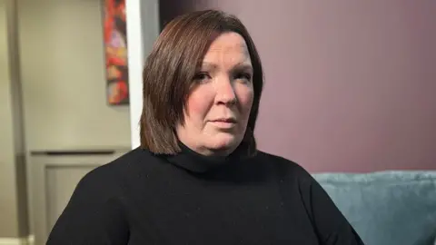 Catriona is wearing a black turtleneck sweater is seated indoors on a light blue couch. The background features a mauve-colored wall with a colorful abstract painting partially visible on the left side, and a doorway leading to another room. The lighting is soft and even, creating a calm atmosphere.