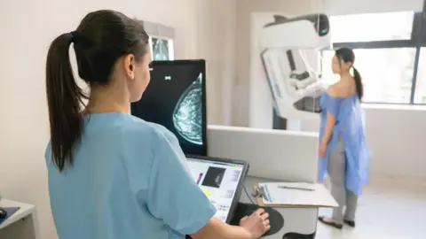 Getty Images A doctor wearing blue scrubs is checking a scan while a woman is standing in the background wearing blue clothing protection receiving a scan.