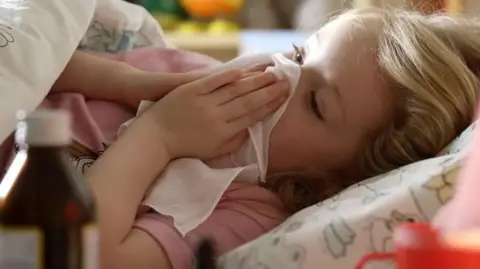 Getty Images File picture of a young girl with long blonde hair with her head on a pillow holding a handkerchief to her nose. She is wearing a pink T-shirt and there is a quilt over her.  