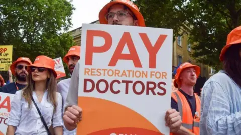 Getty Images Striking doctors stand on a picket line holding orange placards saying Pay Restoration for Doctors, while wearing orange BMA hats