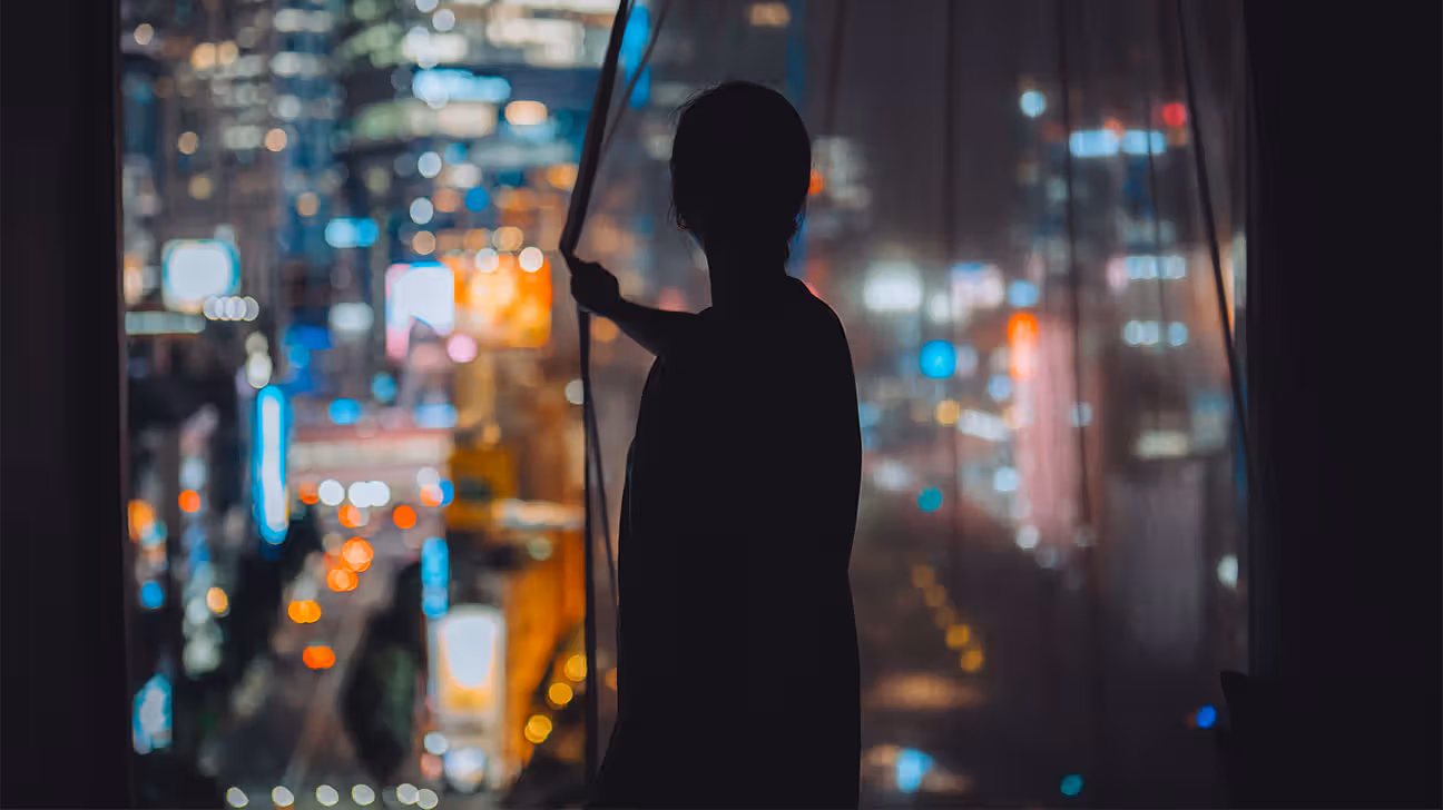 Silhouette of a person in hotel room at night looking out window at bright city lights