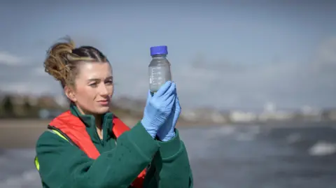 Getty Images A woman with blonde hair tied up in a bun, wearing a green fleece and red lifejacket, holds up a laboratory storage bottle containing water. She is wearing blue latex gloves and examining the water, the background is blurred but shows a river landscape and city surrounding.