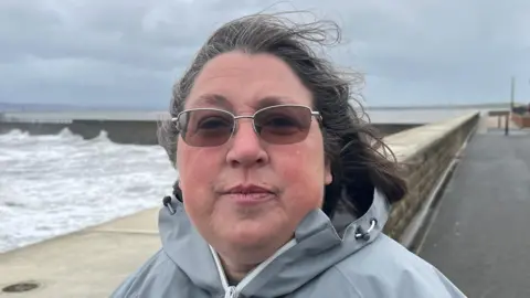 BBC Dawn Lord has light brown sunglasses on with her hair being blown back in the wind. She is wearing a light grey coat with the sea splashing against a sea wall in the background.