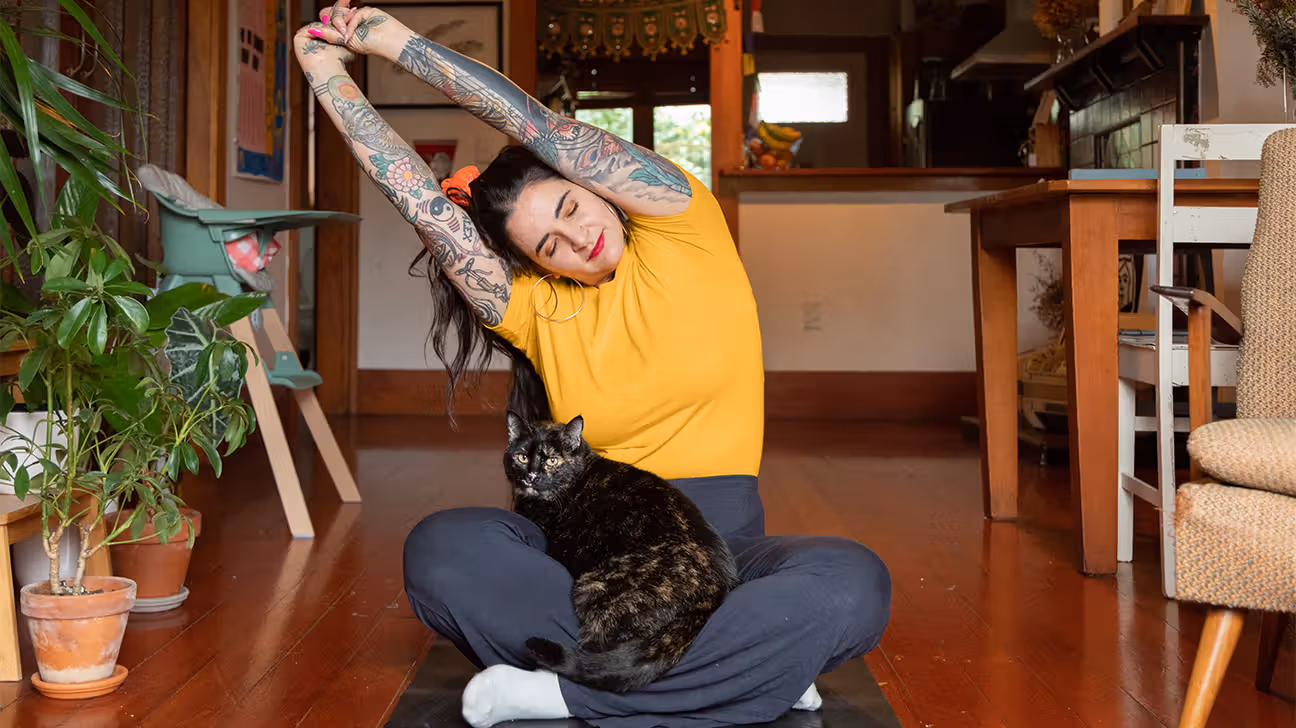 younger white woman with tattoos stretching while in lotus pose on the floor, cat in her lap
