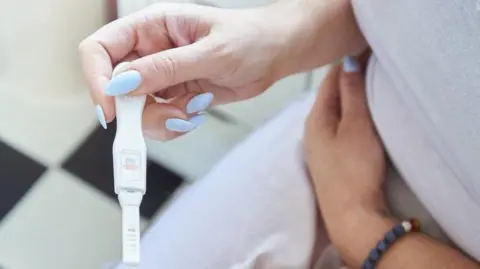 Getty Images Woman with long, light blue fingernails holding a positive pregnancy test while sitting in a bathroom