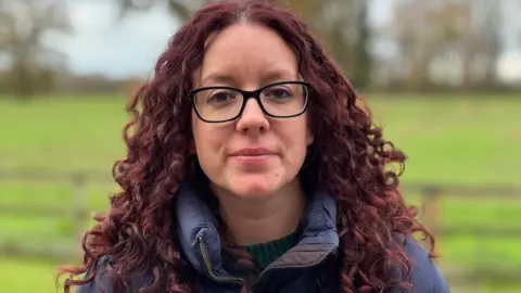 BBC A head and shoulders shot of Kayleigh Griffiths standing in her garden looking into camera with a neutral expression. She has long reddish-brown curlyy hair and is wearing a thick navy jacket. She has black framed glasses.