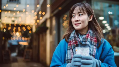 Getty Images Stock photo shows a woman holding a takeaway cup of hot drink wearing blue jumper, gloves and a scarf with Christmas-like street lights in the background.
