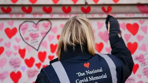Getty Images A woman paints a heart on a wall as part of the Covid memorial. The grey wall is covered with pink and red hearts. The woman has blonde shoulder-length hair and is wearing a dark coat with a dark blue vest over the top which says 
