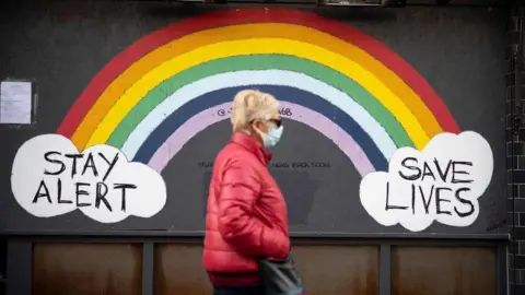 Getty Images A woman wearing a face mask, sunglasses and a red coat walks past a rainbow mural on a wall which reads 