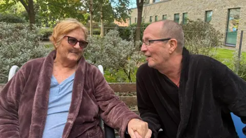 Clare Brixey Woman in purple dressing gown and light blue top. She is wearing sunglasses and holding hands with a man in a black dressing gown looking back at her.