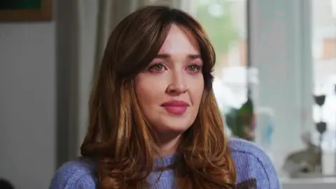 BBC Kate Driver, with long, wavy auburn hair wearing a light purple knit sweater is seated indoors. The background shows a bright window with sheer curtains, some greenery outside, and a few decorative items on the windowsill, including a small plant and a candle holder.