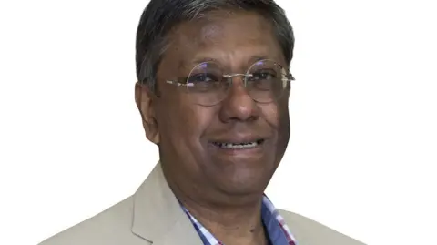 Clatterbridge Cancer Centre A man with dark hair and wire framed glasses with a blue shirt and pale jacket against a plain white background