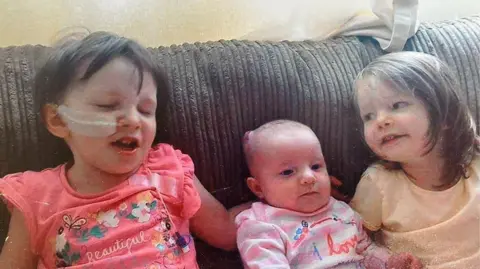 Sam Tucker Two little girls and one baby are sat on a brown sofa, the baby is in the middle. The child on the left has brown hair and a feeding tube in her nose. The child on the right has longer brown hair. 
