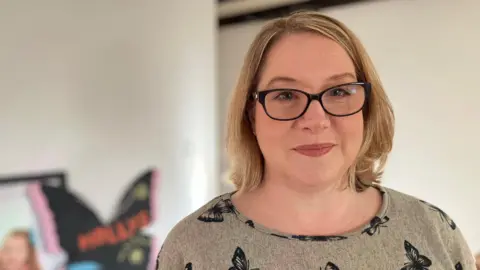 A women is looking at the camera, the photo is just head and shoulders. The women has shoulder length blonde hair and black framed glasses. She is wearing a light brown top with black butterflies on it. 