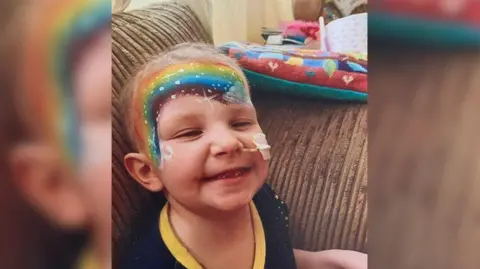 Sam Tucker A child who is undergoing treatment is smiling at the camera. She has a brightly coloured rainbow painted on her face. 