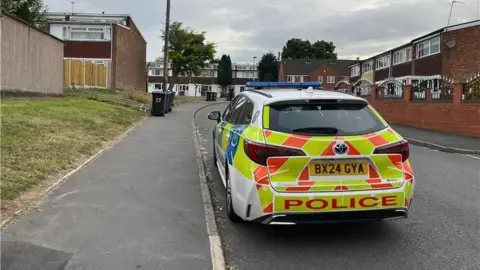 BBC/Louise Brierley A police car is parked on a road in a residential area. There are houses on the right and left