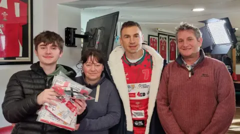 Family handout Kevin Sinfield stands shoulder-to-shoulder with Kyle's parents, and brother, Liam, at a rugby club. Wales rugby jerseys are hung on the wall behind them.