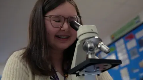 Girl with long brown hair and wearing glasses stairs down a microscope in a biology class.