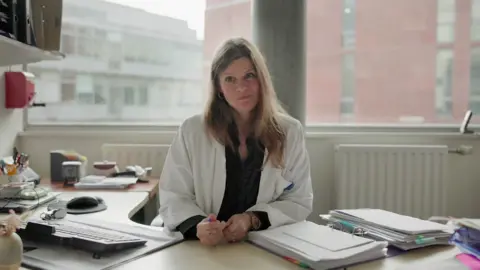 Dr Kasper sits at her work desk, wearing a white coat