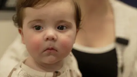 Oscar, aged five months, sits in his mother's arms. He has brown hair with a curl and is wearing a white and brown outfit.