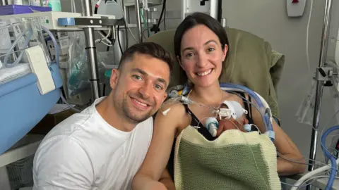 PA Media A dark haired young man sits in hospital with a dark haired young woman. The woman hold a small baby on her chest. The baby is wired to medical equipment.