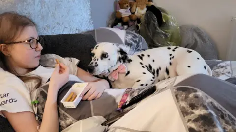 Cancer Research UK handout Sophie Claxton with long brown hair lying on the sofa under a quilt eating bread sticks and soft cheese snack while her white and black spotted dalmatian dog, Bonnie, lies next to her staring at the snack.  Bonnie is wearing a pink white and black bow tie collar.