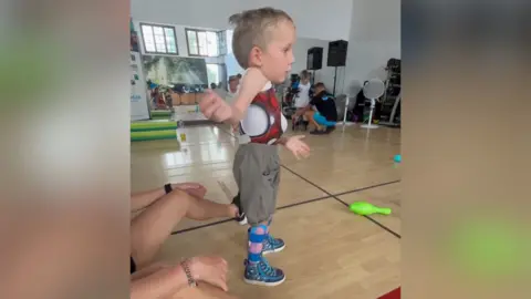 Contributed Edward in a physiotherapy room, stood upright. He is wearing a body brace, a white vest, dark jogging bottoms and blue boots. An adult is sitting behind him.