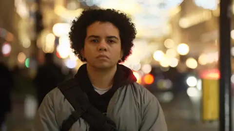BBC Keira Bell stands in front of a dark, black curtain, wearing a black hoodie with red and yellow designs near the elbows. She has dark, curly, short hair, dark brown eyes and wears earrings in her ears.