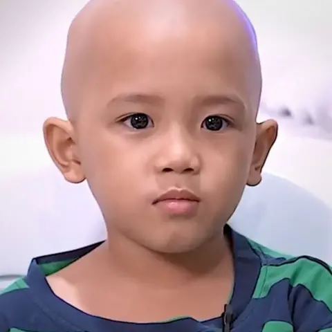 Chance Letikva Khalil, a little Filipino boy, is wearing a green and blue striped t-shirt, has a shaved head, and has a small microphone clipped to his top. There is a white hospital background. He faces the camera, mid-speech.