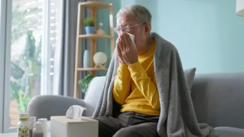 Getty Images An older woman with grey hair and glasses blows her nose while sitting on a sofa with a grey blanket round her shoulders. There is a box of tissues on the table in front of her as well as various bottles that may be medicine. 