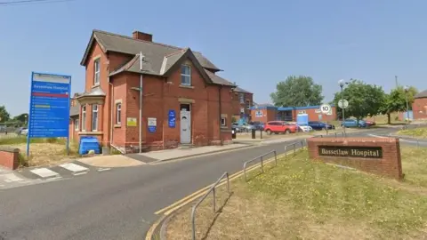 Google Google streetview image of the entrance to Bassetlaw hospital, showing a detached Victorian red brick house next to a name plate, with cars parked in the background
