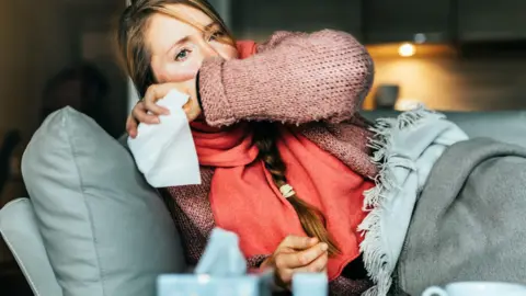 Getty Images An unwell woman sits on her sofa, coughing 