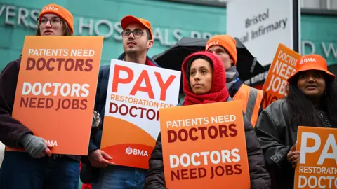Getty Images People holding placards saying 'patients need doctors, doctors need jobs' stand outside wearing hats and thick clothes outside Bristol Royal Infirmary on Wednesday.