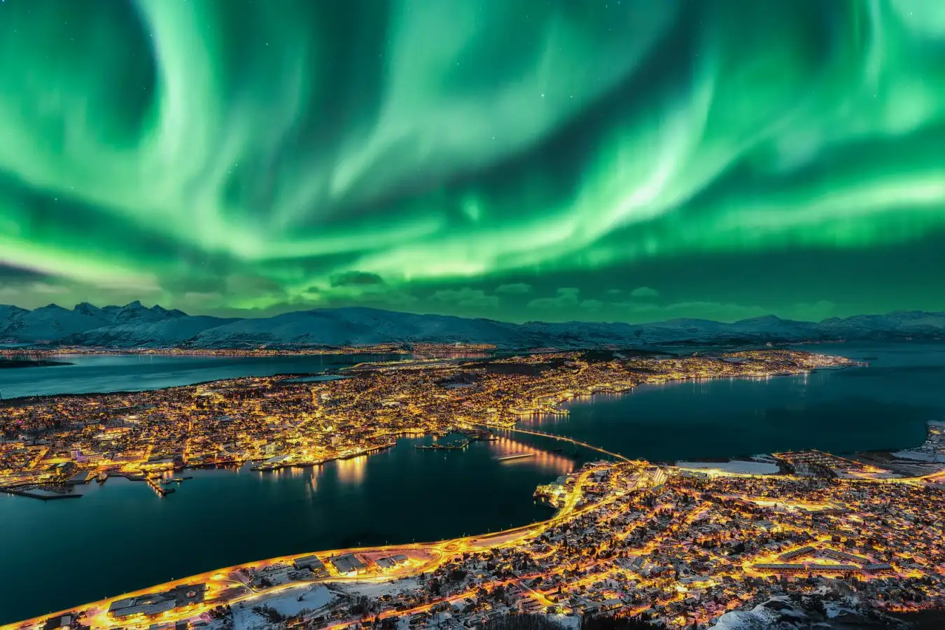 Aurora Borealis dancing over Tromso Urban Skyline. Tromso Panorama and winter night landscape with Northern Lights (Aurora Polaris) on a cold night, with starry sky, polar lights and snowcapped mountains on Kvaloya Island in the background. Cityscape with Brightly Street Light of the Arctic Capital, Northern Norway