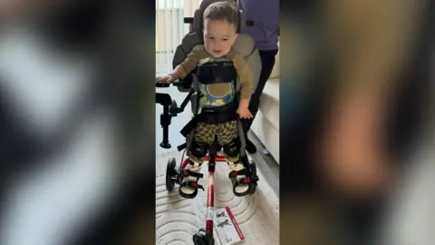 Samantha Williams Lucian is standing on an assisted piece of kit inside a room. The appliance has his torso, hips, knees and feet wrapped so he is able to stand up straight.