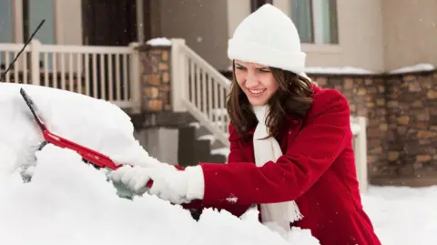 Getty Images A young woman wearing a long red coat and white hat, scarf and gloves, scrapes a thick layer of snow off the windscreen of her car. She is parked outside her house. 